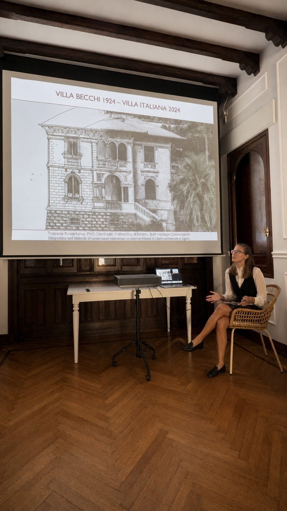 A speaker presenting on the villa’s history beside a projector screen in a wood-paneled room with herringbone parquet floors and beamed ceiling