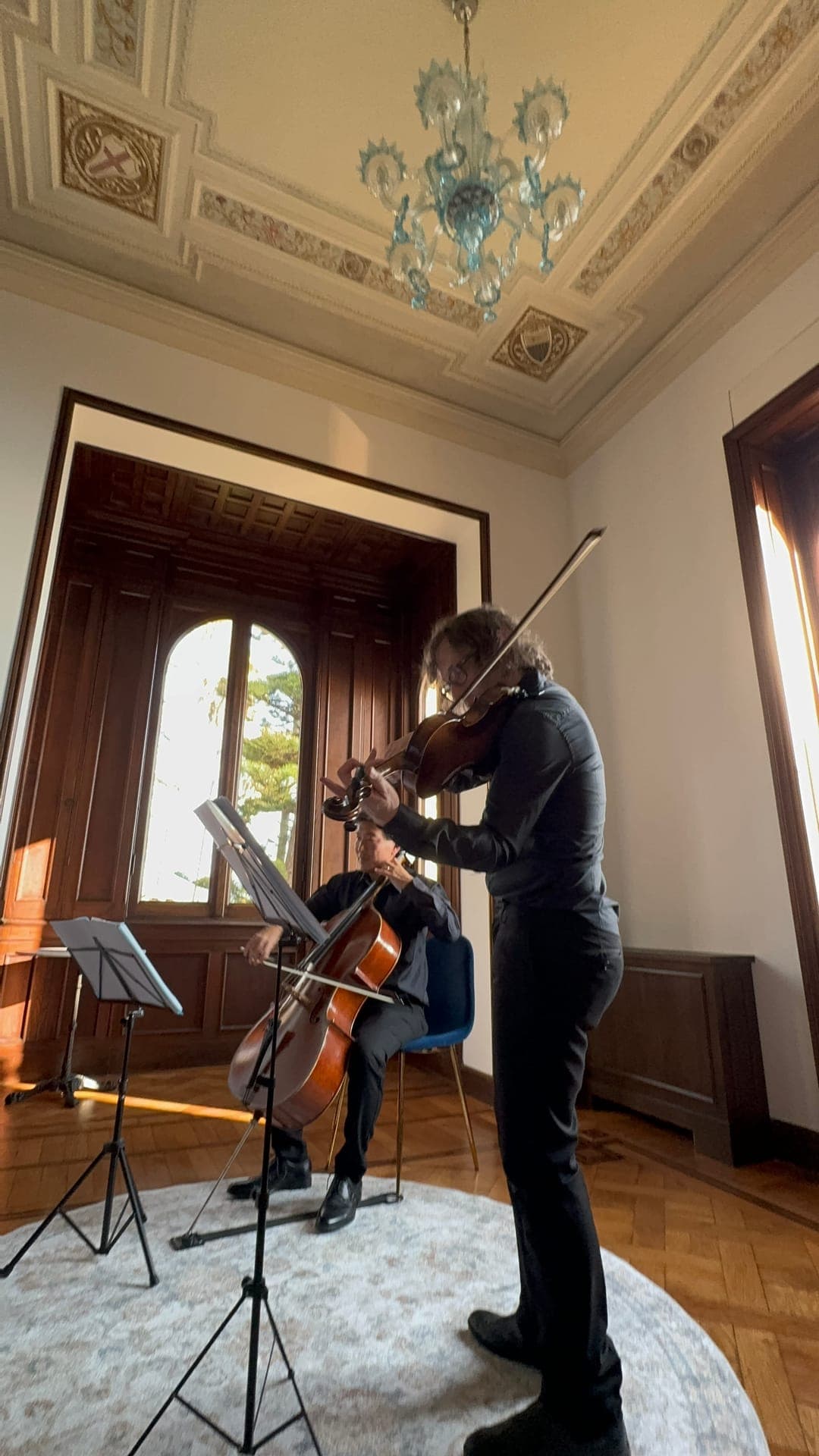 A violinist and cellist performing in an ornate salon with a Murano glass chandelier and frescoed ceiling, before an arched window with a sea view
