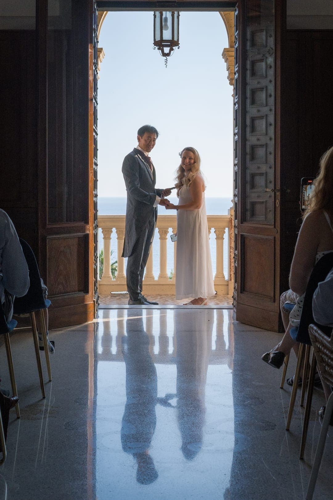 A couple exchanging vows framed in a grand doorway, silhouetted against the sea and a balustraded terrace, with guests seated on either side