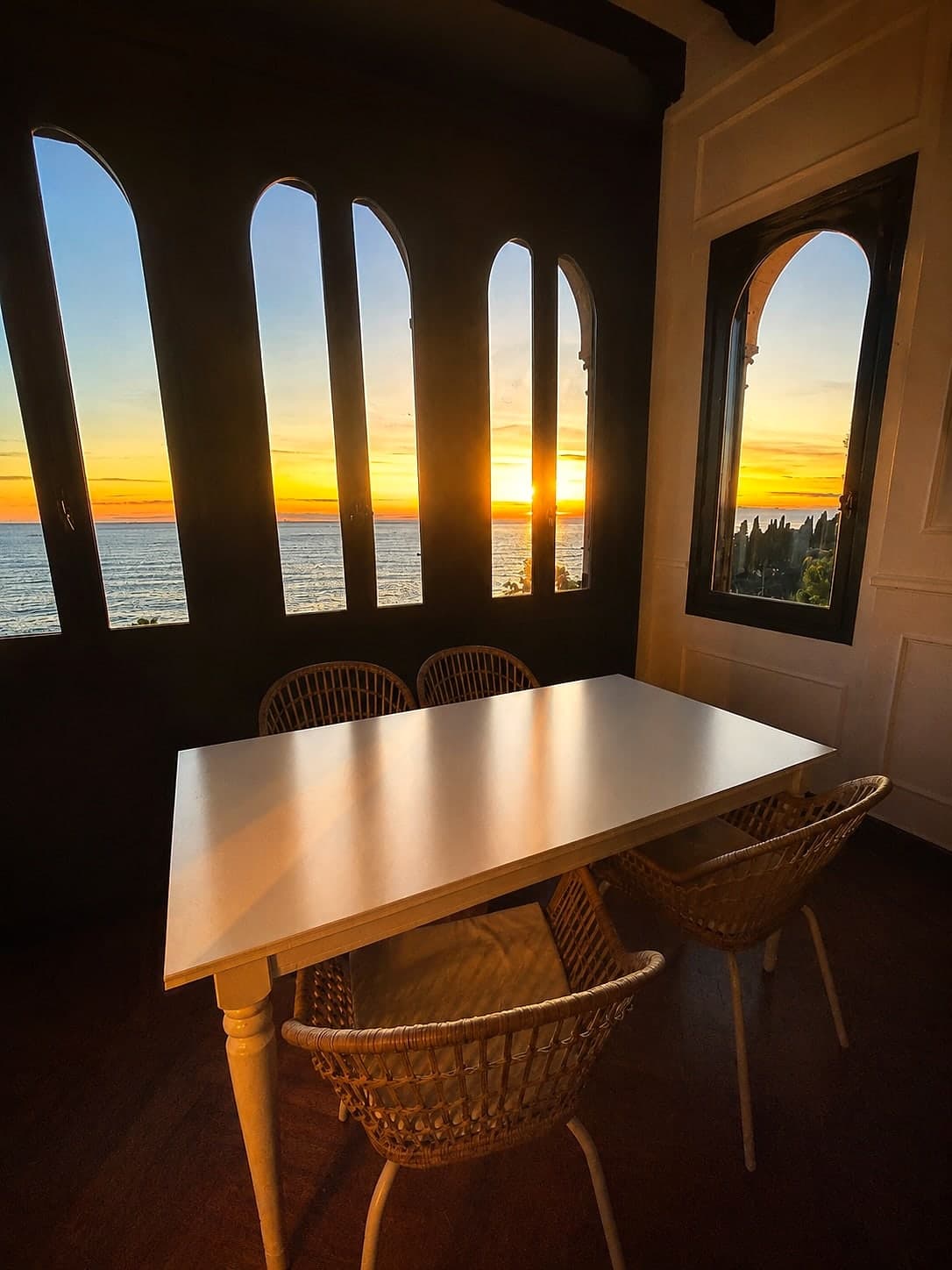 Dining nook with woven rattan chairs framed by the villa's tall arched windows overlooking a glowing sunset over the Mediterranean
